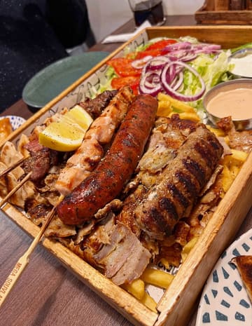 A top-down, close-up shot of a large Greek mixed grill platter served on a wooden board, featuring a variety of meats including skewers, sausages, and gyros, accompanied by chips, salad, and dips.