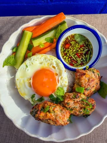 A vibrant, overhead shot of a homemade lemongrass chicken meatball bowl, featuring a perfectly cooked fried egg, fresh cucumber and carrot matchsticks, and a side of dipping sauce, presented on a simple white plate.