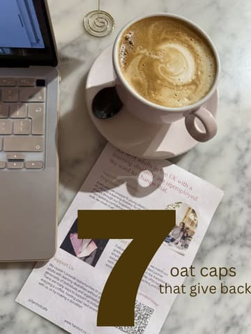 A clean, well-composed flat lay photograph featuring a cup of coffee on a marble table alongside a laptop and a flyer for a social enterprise cafe. The image effectively communicates a 'work from cafe' vibe while highlighting the charitable aspect of the featured coffee shops.