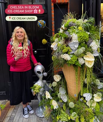 A photo of a person holding an ice cream cone while standing next to a large floral display (Chelsea in Bloom) and an alien statue outside an ice cream shop.