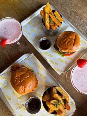 A bright, overhead flat-lay shot of a bottomless brunch spread at The Canary, featuring two burgers, two portions of fries, and two cocktails on branded paper.