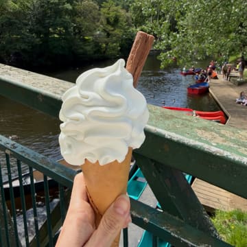 A handheld shot of a soft-serve ice cream cone with a chocolate flake, held against a backdrop of a river with boats, likely taken during a casual day out.