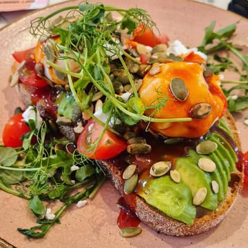 A close-up, high-angle shot of avocado and feta toast topped with poached eggs, sweet chilli sauce, and pumpkin seeds, served at a play cafe.