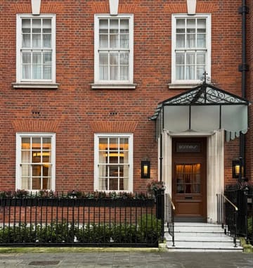 A professional, eye-level architectural photograph of the exterior of Bonheur restaurant in Mayfair. The image captures the classic red brick facade, symmetrical windows, and the elegant glass canopy entrance, conveying a sense of prestige and high-end hospitality.