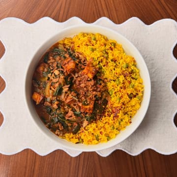 A top-down, well-composed photograph of a homemade paneer curry served alongside vibrant yellow pea rice in a white bowl, set on a scalloped placemat on a wooden surface.