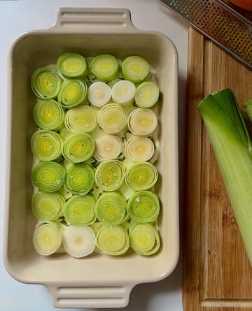 A clean, overhead food photography shot featuring sliced leeks arranged neatly in a baking dish, likely in preparation for a roasted vegetable recipe. The lighting is soft and natural, highlighting the texture and vibrant green tones of the produce, while the composition is balanced and professional.