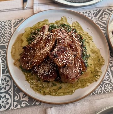 A top-down, overhead shot of a home-cooked meal featuring grilled lamb chops topped with sesame seeds, served over a vibrant green salsa verde. The dish is presented on a ceramic plate set against a patterned placemat, capturing a rustic and appealing dining aesthetic.