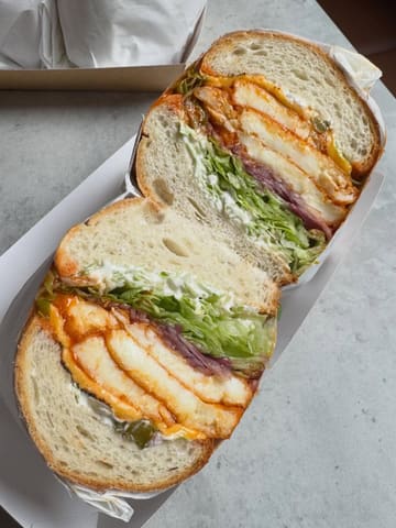A close-up, top-down shot of a halloumi sandwich cut in half, showcasing the layers of buffalo-dipped halloumi, lettuce, and sauce. The image is bright and clear, highlighting the texture of the bread and the freshness of the ingredients.