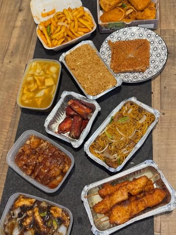 A top-down, flat-lay photograph of a large Chinese takeaway spread arranged on a dark grey surface, featuring various dishes in plastic and foil containers.