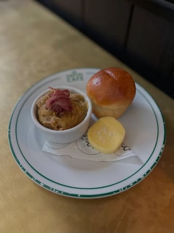 A casual, top-down shot of a small plate featuring pease pudding with ham hock, a milk loaf, and a slice of butter. The image is well-lit and clear, capturing the food in a straightforward manner without professional styling.