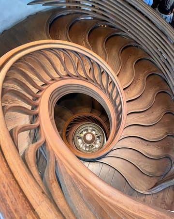 A high-angle, symmetrical shot looking down a striking wooden spiral staircase at a high-end restaurant. The image focuses on the architectural detail and the central light fixture, creating a visually compelling and sophisticated aesthetic.