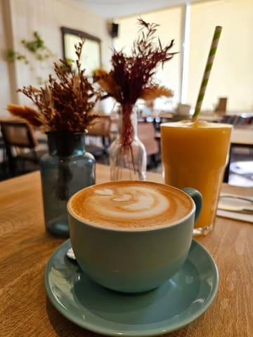 A casual, cozy shot of a coffee and a juice at a cafe, featuring latte art and dried flower decor.