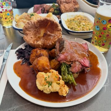 A high-angle shot of a Sunday roast dinner featuring sliced beef, a large Yorkshire pudding, roast potatoes, cauliflower, and red cabbage, served in a pub setting with beer glasses visible.