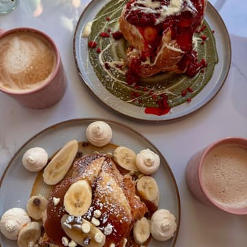 A bright, overhead flat-lay shot featuring two plates of brioche French toast topped with fruit and cream, accompanied by two cups of coffee on a white marble surface.