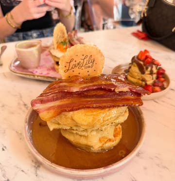 A close-up, eye-level shot of a stack of pancakes topped with bacon, served on a marble table in a cafe setting. The image captures a casual dining moment with other plates and personal items visible in the background.