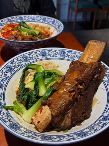 A close-up, high-angle shot of a bowl of braised short rib biang biang noodles, with a second bowl of noodles visible in the background.