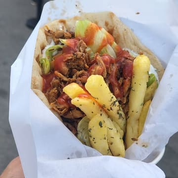 A close-up, eye-level shot of a chicken gyros served in a paper wrapper, featuring fries, lettuce, and sauce, captured at a street food stall.