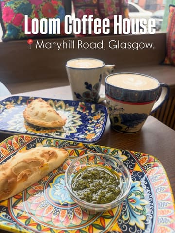 A high-angle shot of a table at Loom Coffee House featuring two empanadas, a bowl of chimichurri, and two coffees in patterned mugs. The image captures a casual, cozy cafe setting with natural window lighting.