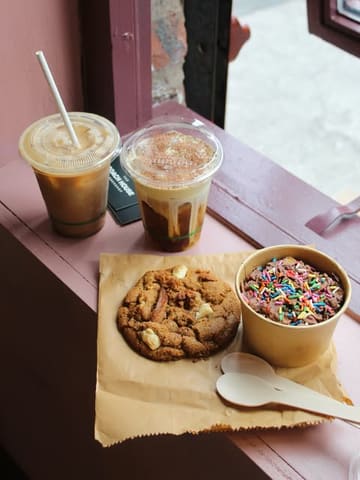 A high-angle, natural-light shot of a coffee shop spread featuring two iced coffees, a large cookie, and a small chocolate dessert pot, arranged on a rustic brown paper surface.