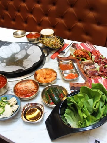 A high-angle, overhead shot of a Korean BBQ spread featuring various banchan, raw marinated meats, lettuce wraps, and a grill. The table is well-laid out, showcasing the variety of the meal.