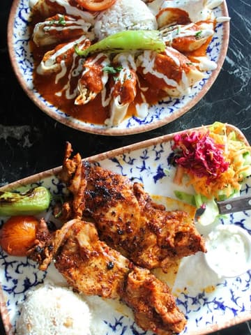 A high-quality, overhead flat-lay photograph of two Turkish dishes served on patterned plates against a dark marble table. The bottom plate features grilled chicken, rice, salad, and grilled vegetables, while the top plate shows a dish with tomato sauce, yogurt, and rice.