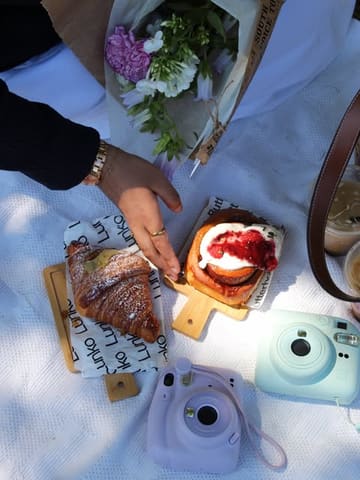 A picnic-style flat lay featuring a croissant and a raspberry cinnamon bun, accompanied by iced lattes and flowers, capturing a relaxed outdoor dining aesthetic.