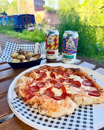 A high-quality, appetizing shot of a pepperoni pizza and garlic doughballs paired with craft beer, set against a scenic canal-side backdrop.