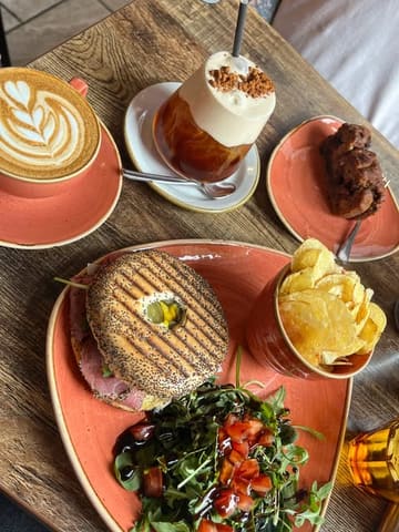 A bright, natural-light flat lay of a brunch spread featuring a bagel, salad, chips, a latte, and an iced coffee. The image captures a casual, inviting cafe atmosphere.