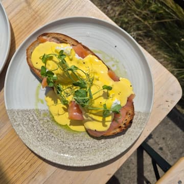 A top-down shot of an Eggs Royale dish served on a ceramic plate, placed on a wooden table outdoors. The dish features poached eggs, smoked salmon, and hollandaise sauce garnished with pea shoots on toasted sourdough.