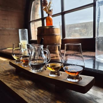 A well-composed, atmospheric shot of a whisky tasting flight served on a wooden board, positioned on a window ledge in a rustic bar setting.