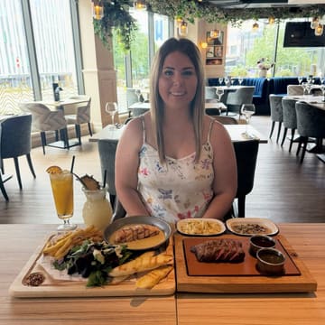 A promotional image featuring a person sitting at a restaurant table with a spread of food including a salmon fillet, steak, various sides, and two cocktails.