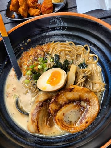 A high-quality, top-down food photography shot of a bowl of black garlic tonkotsu ramen, featuring a perfectly soft-boiled egg and seared pork belly, with a side dish of karaage visible at the top of the frame.