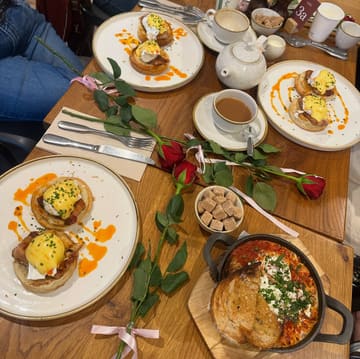 A top-down flat lay photograph of a brunch spread featuring plates of eggs benedict served on croissant swirls, a skillet dish with toasted bread, and hot drinks, with red roses arranged diagonally across the table.