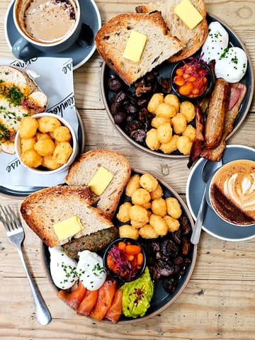 A high-quality, overhead flat-lay shot of a brunch spread featuring two main plates, two coffees, and a side of tater tots. The lighting is bright and natural, highlighting the textures of the sourdough, poached eggs, and various breakfast components.
