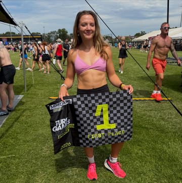 A photo of a woman holding a first-place flag at the Turf Games fitness competition, standing on a grass field.