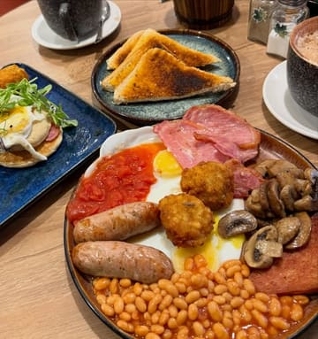 A top-down shot of a hearty cafe breakfast spread, featuring a full English breakfast with spam and a separate plate of pancakes with bacon and eggs.