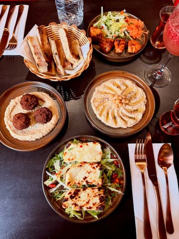 A top-down flat lay photograph of a Turkish meal spread featuring hummus, falafel, grilled halloumi, mixed grill, and bread, arranged on a black table.