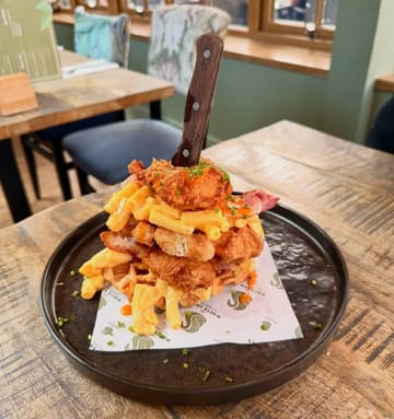 A high-angle, close-up shot of a towering comfort food stack featuring fried chicken, waffles, bacon, and mac and cheese, served on a branded paper-lined plate with a knife inserted into the top.
