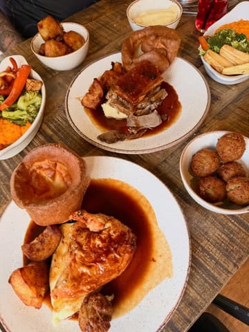 A top-down, flat-lay photograph of a Sunday roast spread at a pub, featuring multiple plates including roast chicken, a trio of meats, roast potatoes, mash, and vegetables.