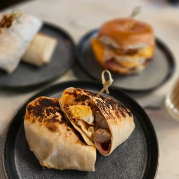 A well-composed, appetizing shot of a breakfast burrito cut in half, served on a dark plate, with a blurred background showing other menu items. The lighting is natural and flattering, highlighting the texture of the toasted tortilla and the filling.