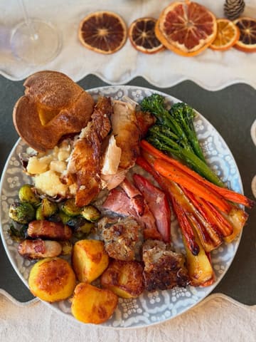 A top-down, well-lit photograph of a traditional British Christmas roast dinner, featuring roast chicken, potatoes, pigs in blankets, stuffing, and vegetables. The image is captured in a home-cooking style with thematic dried orange decorations, presenting a warm and festive atmosphere.