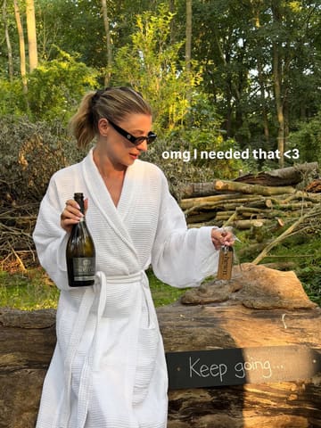 A woman in a white bathrobe sits on a log in a wooded area, holding a bottle of Prosecco and a wine glass with a branded tag, promoting a relaxing countryside retreat experience.