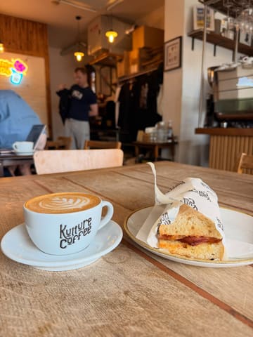 A casual, well-lit shot of a coffee and a bacon sandwich on a wooden table at a cafe, showcasing the venue's branding on the cup and packaging.