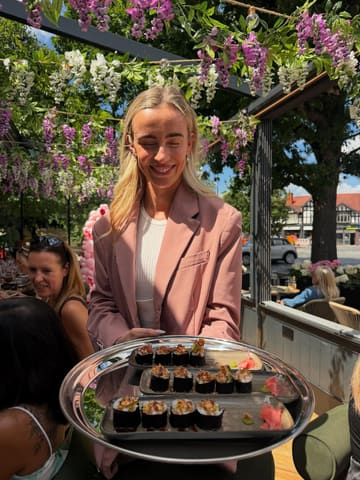 A portrait-style shot of a woman holding a tray of sushi rolls on an outdoor terrace, with wisteria-like flowers overhead. The image captures a moment from a bottomless brunch, showcasing the venue's outdoor seating and atmosphere.