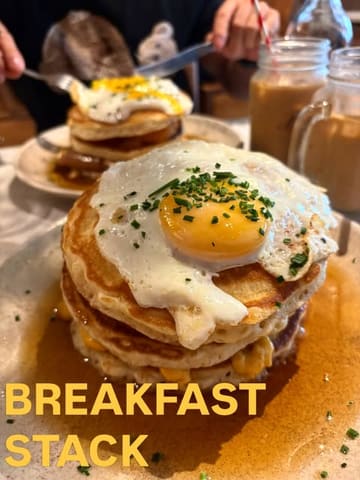 A close-up shot of a breakfast pancake stack topped with a fried egg, served at a cafe. The image captures the texture of the pancakes and the runny yolk, presented in a casual dining setting.