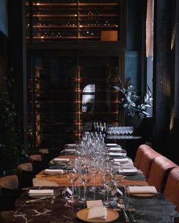 A sophisticated, moody shot of a long, elegantly laid dining table in a high-end restaurant, featuring a wine cellar in the background.