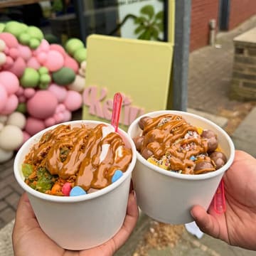 A bright, high-quality close-up shot of two cups of frozen yogurt held in hands, showcasing generous toppings and sauces. The background features a branded sign and a balloon arch, providing a clear, inviting context for the venue.