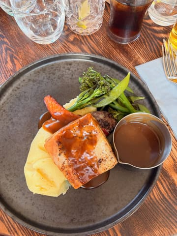 A top-down, natural light shot of a pork belly main course served with mashed potatoes, tenderstem broccoli, and a side of gravy, presented on a dark plate on a wooden table.