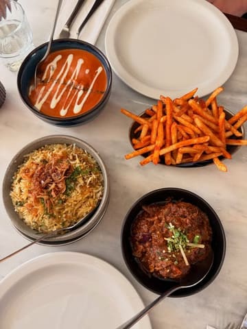 A bright, overhead flat-lay photograph of an Indian meal spread, featuring bowls of curry, rice, and masala fries arranged on a clean marble surface. The natural lighting enhances the vibrant colours and textures of the food, making for an inviting and appetising composition.