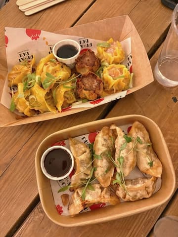 A top-down, overhead shot of two takeaway containers filled with dumplings and gyoza, served with dipping sauce, placed on a wooden table.